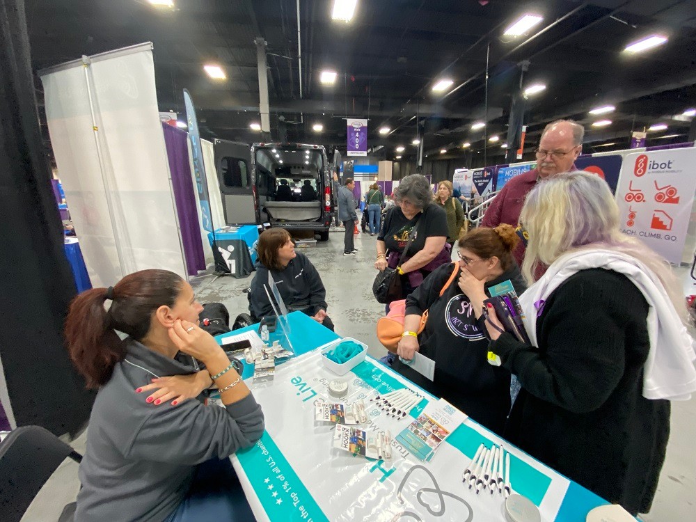 In a photo taken from just past Kelly Green's shoulder, Kelly is seated at the teal-themed Help Hope Live booth at Abilities Expo NY Metro 2024 listening to client Amy Sherwood talk to a family that has stopped by the booth. Kelly has light skin, red-brown hair pulled into a ponytail, and a gray sweater with bright magenta nails. Seated in her wheelchair, Amy Sherwood has light skin and shoulder-length straight brown hair with bangs, and her black labrador service dog Dolly Pawton is within reach. She speaks with three women and one man with Help Hope Live brochures and pens visible on the booth table just in front of them.