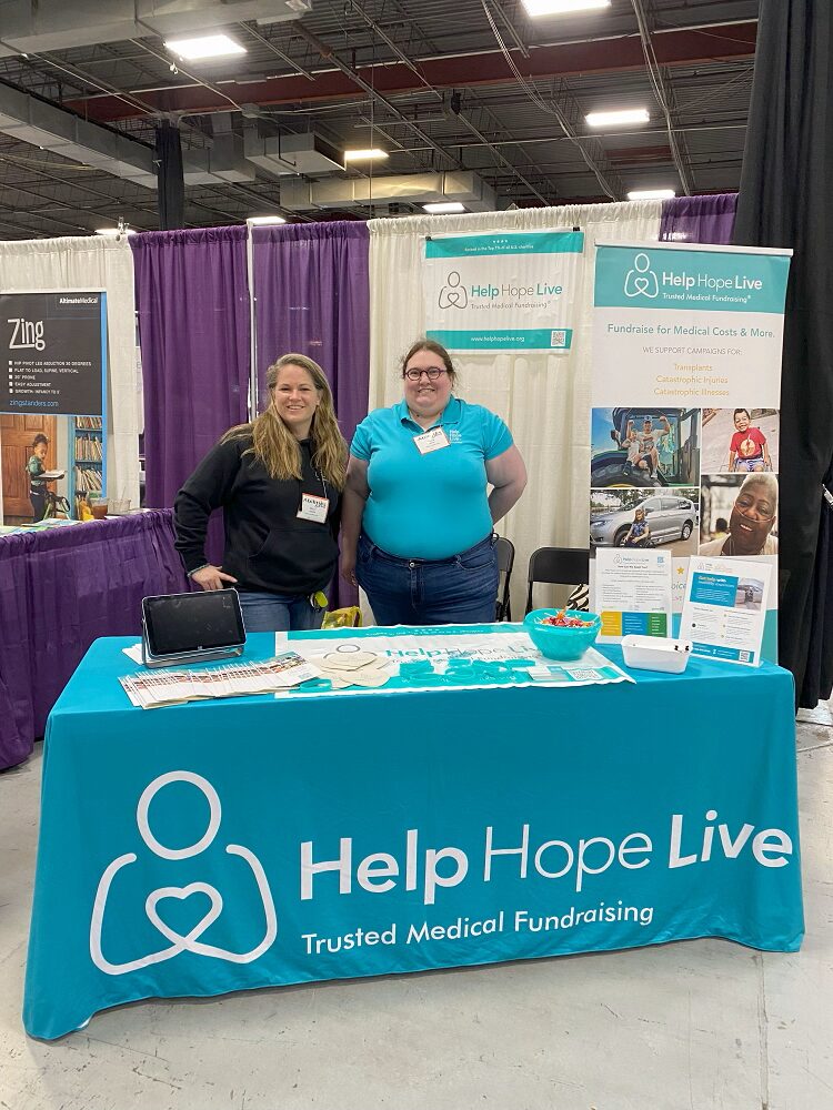 Stacia and Sonny from Help Hope Live stand behind the teal Help Hope Live: Trusted Medical Fundraising booth at Abilities Expo NY Metro 2024. Stacia has light skin, round purple glasses, and curly brown hair and she wears jeans and a teal Help Hope Live polo shirt. Sonny has light skin, golden-brown straight hair, a black Help Hope Live Nike fleece, and jeans. The table features pens, signs, teal bracelets, and photos of Help Hope Live clients.