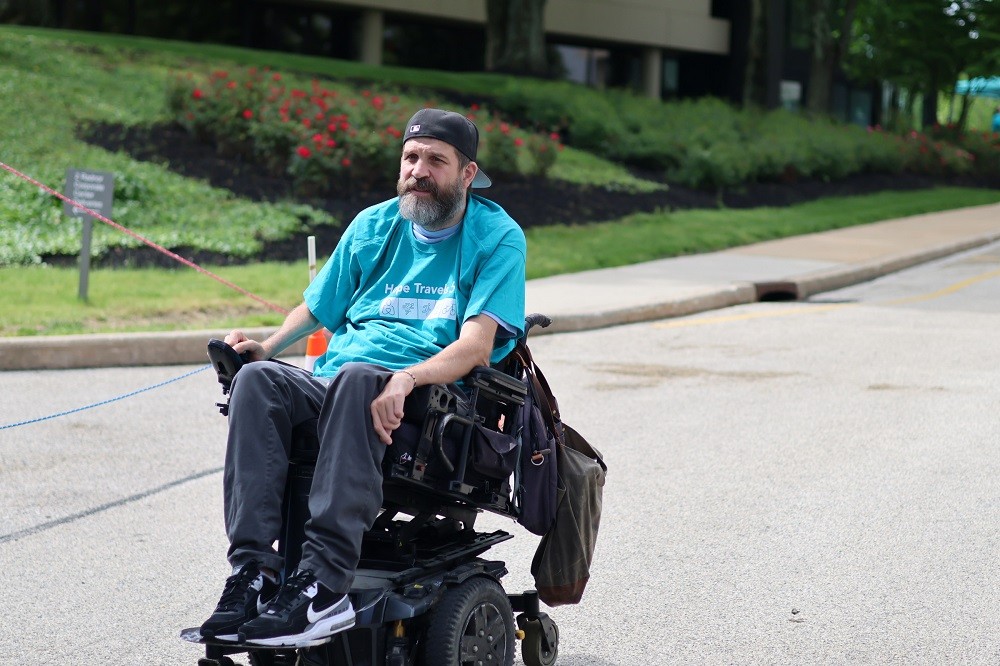 Help Hope Live client Ambassador Jeff Harris sports a teal Hope Travels t-shirt and sneakers with a backwards ball cap as he rolls down the street in his black power chair at Hope Travels 2024. He has light skin and a gray and brown beard.