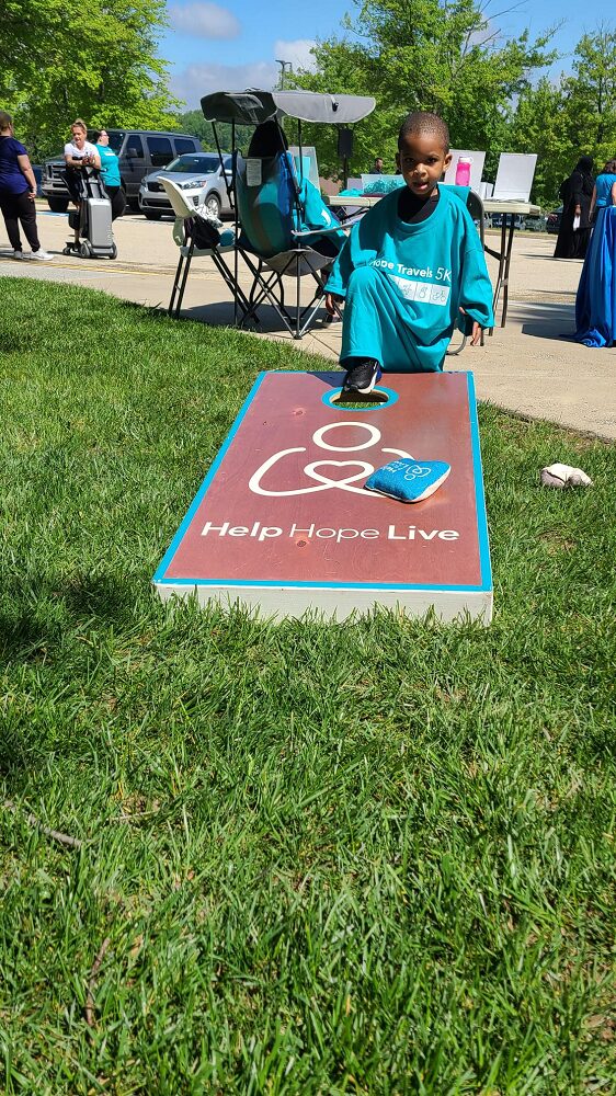 A child with brown skin and short dark hair stretches a sneakered foot onto a Help Hope Live branded cornhole set. He is swimming in an oversized teal Hope Travels 5K t-shirt outdoors at the Hope Travels 2024 event.