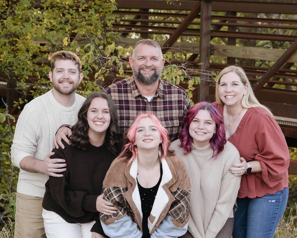 A family portrait outdoors of kidney transplant recipient Josh Raymond with his wife, an adult son, an adult daughter, and two young adult daughters. Everyone is dressed in fall clothing such as turtlenecks in neutral color shades. Josh has light skin, striking blue eyes, short gray hair, and a gray beard. His wife has light skin, light eyes, long blonde hair, and gold hoop earrings. His son has light skin, red hair and a red beard, and blue eyes. His adult daughter has light skin, brown eyes, and shoulder-length wavy brown hair. His two young adult daughters both have light skin and hazel eyes with shoulder length wavy hair - one has brown hair streaked with bright salmon pink, and the other has mulberry purple hair.