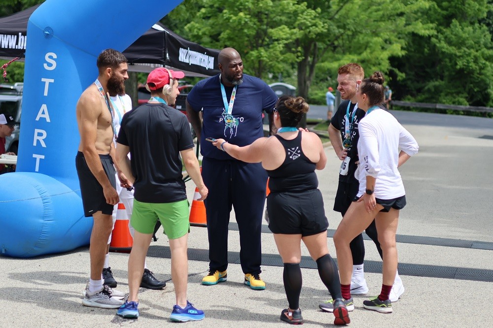 At the finish line of Hope Travels 2024 outdoors, Barrett Brooks speaks to six young adult runners in athletic gear and sneakers. Barrett has brown skin and a short black and gray goatee and he wears a Hope Travels medal along with several of the runners.