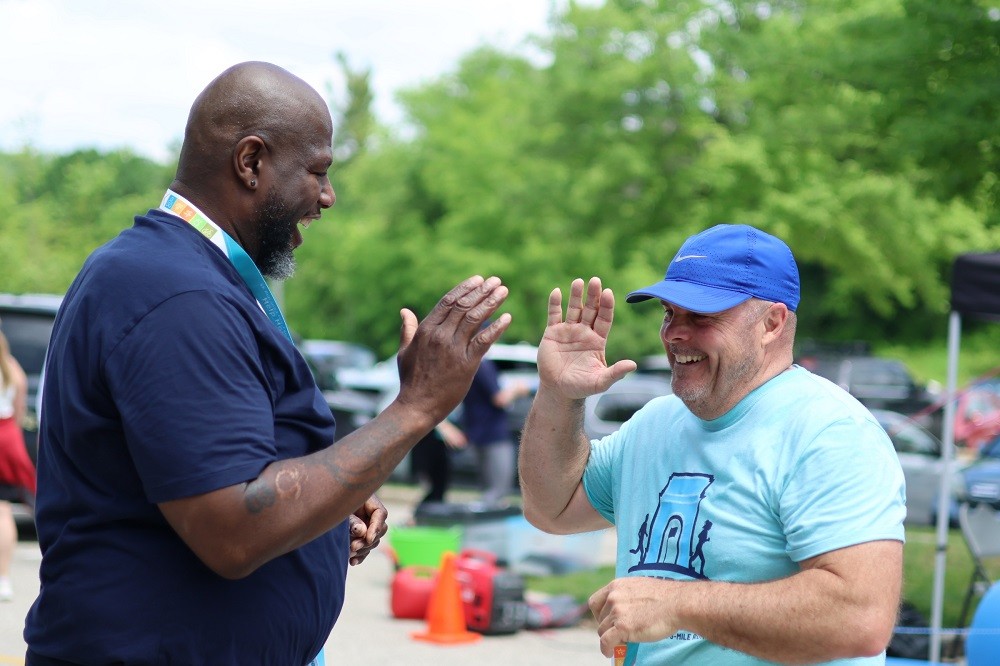 A runner finishes Hope Travels 5K in 2024 and reaches to give Barrett Brooks a high five. The runner has light skin, a light blue t-shirt, and a blue Nike ball cap. Barrett has brown skin and a short black and gray goatee. Both are grinning.