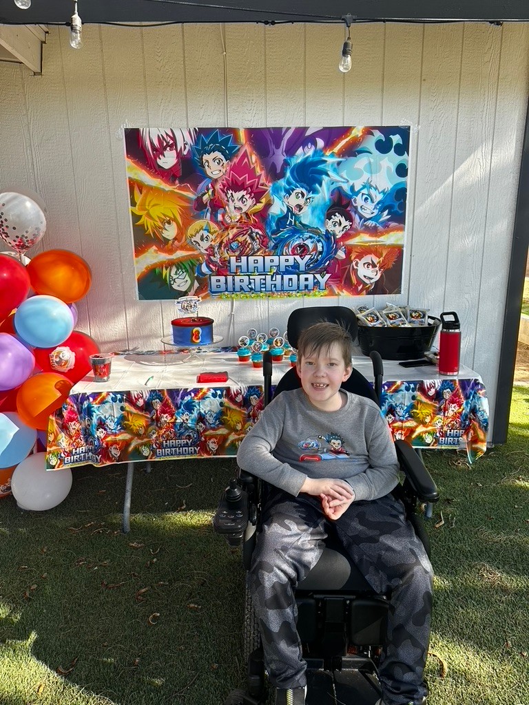 Carson smiles in front of a special outdoor setup for his birthday featuring anime character posters, balloons, a cake, and favors. Carson has light skin, brown hair with wispy bangs, a gray long-sleeved anime shirt, and gray and black camo sweatpants. He is seated in a black power chair.