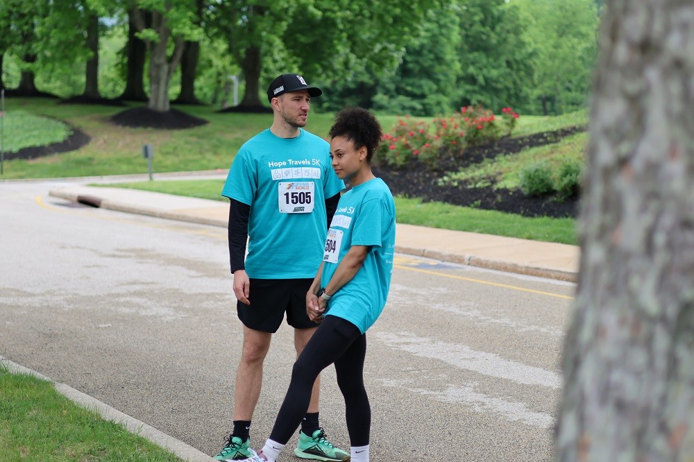 Two participants in Hope Travels 2024 stand outdoors in athletic gear and matching Hope Travels 5K teal t-shirts. The young man has light skin, black shorts, and a black fitted cap. The young woman has brown skin, curly black hair pulled back, and black leggings.