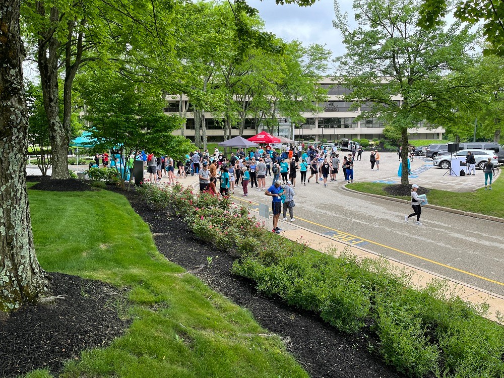 Dozens of participants congregate in a corporate center parking lot at Hope Travels 2024. They wear athletic gear with some sporting runner numbers on their shirts. Surrounding them is spring greenery with manicured grass and green trees.