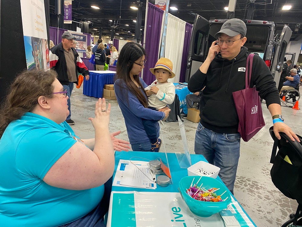 Stacia performs sign language with her two hands as she sits behind the teal Help Hope Live booth at Abilities Expo New York Metro 2024 speaking to a man wearing a ball cap with glasses beside a woman with straight black-brown hair holding a young child in a straw hat. Stacia has light skin, round purple glasses, and curly brown hair and she wears jeans and a teal Help Hope Live polo shirt.