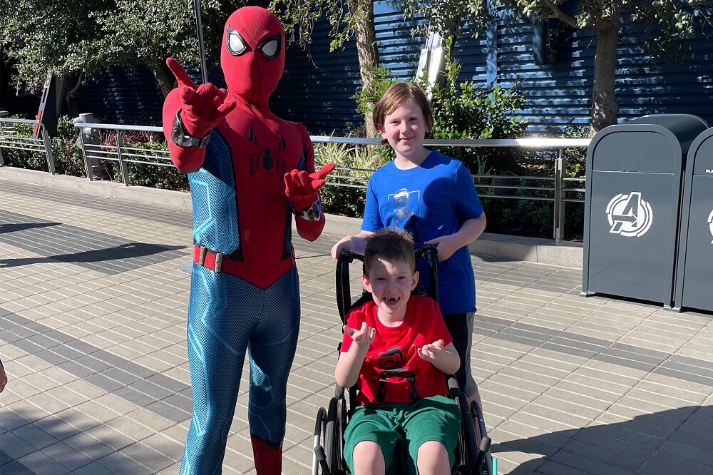 On an outdoor walkway, Carson and his older brother pose next to someone dressed in an elaborate blue and red Spider-man superhero costume. Carson is seated in a black manual wheelchair making spidey-web-shooting hands to match the costumed hero. He has light skin, short light brown hair, and his two front teeth missing. Carson wears green shorts and a red t-shirt for The Mandalorian. His brother standing behind him is slightly older with a blue Mandalorian t-shirt, light skin, and longer straight light brown hair.