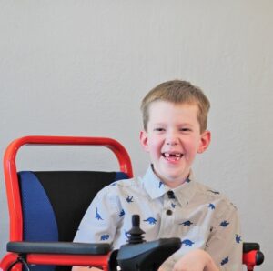 Carson Tussing is seated in a red and blue electric mobility device. He has light skin, his two front teeth missing, and short light brown hair and he wears a collared short-sleeved shirt with dinosaurs printed on it.