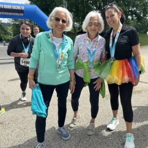 A photo of three women next to the Bryn Mawr Racing Co starting line at Hope Travels 2024. Two of them are significant Help Hope Live supporters: current Help Hope Live Executive Director Kelly L Green, who has light skin, dark hair pulled back, sneakers, a black Help Hope Live polo, and a rainbow tutu – and co-founder Pat Kolff, who has light skin, brown sunglasses, sneakers, and white shoulder-length hair. The three women wear Hope Travels 5K medals around their necks.