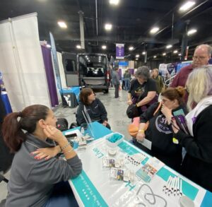 In a photo taken from just past Kelly Green's shoulder, Kelly is seated at the teal-themed Help Hope Live booth at Abilities Expo NY Metro 2024 listening to client Amy Sherwood talk to a family that has stopped by the booth. Kelly has light skin, red-brown hair pulled into a ponytail, and a gray sweater with bright magenta nails. Seated in her wheelchair, Amy Sherwood has light skin and shoulder-length straight brown hair with bangs, and her black labrador service dog Dolly Pawton is within reach. She speaks with three women and one man with Help Hope Live brochures and pens visible on the booth table just in front of them.