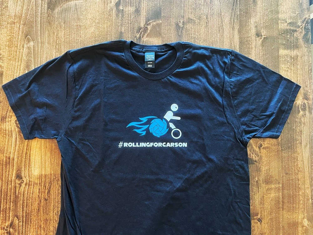 A navy blue t-shirt says #RollingforCarson and depicts a person in a wheelchair leaning forward with a determined competitive attitude. The wheelchair is made up of light blue geometric spikes with blue flames coming from the back.