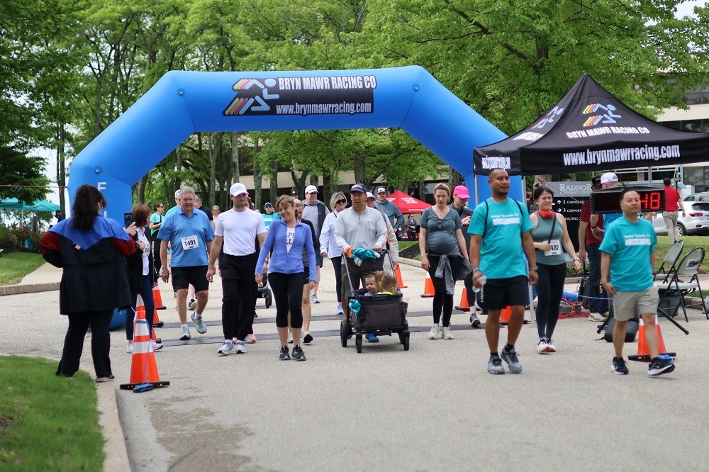 Walkers take off at the Bryn Mawr Racing Co starting line at Hope Travels 2024 outdoors. The walkers are a mix of ethnicities and ages in athletic gear with some wearing teal Hope Travels 5K t-shirts.