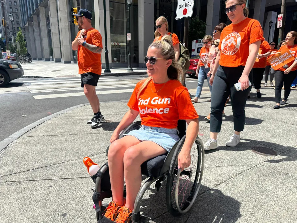 38-year-old Amanda Parezo is seated in her manual wheelchair on a Philadelphia city street wearing orange sneakers and an orange shirt that reads End Gun Violence. She has light skin, blonde hair, and sunglasses. A group of 10 people walking behind her all wear the same t-shirt.