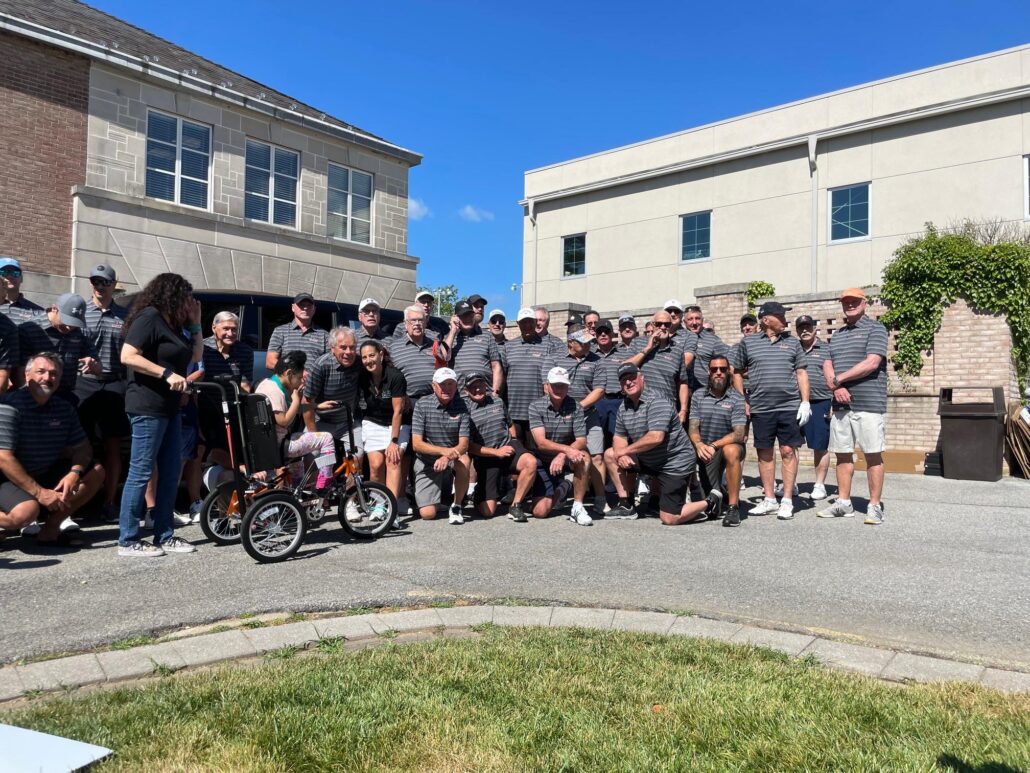 A group of golfers in Flyers Alumni gray polo shirts with Help Hope Live Executive Director Kelly L Green and 8-year-old adaptive bike recipient Josie Johnson.