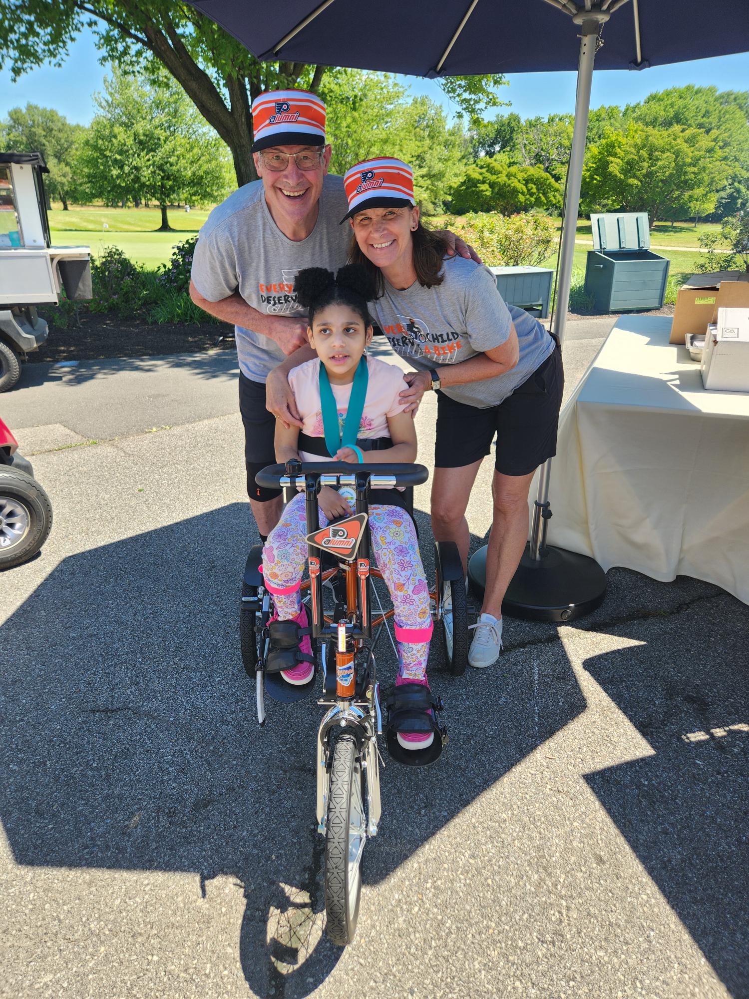 Two Flyers golf outing participants outdoors wearing Every Child Deserves a Bike t-shirts with 8-year-old adaptive bike recipient Josie Johnson who has light brown skin and curly black hair. One of the participants is Flyers alum Brad Marsh.