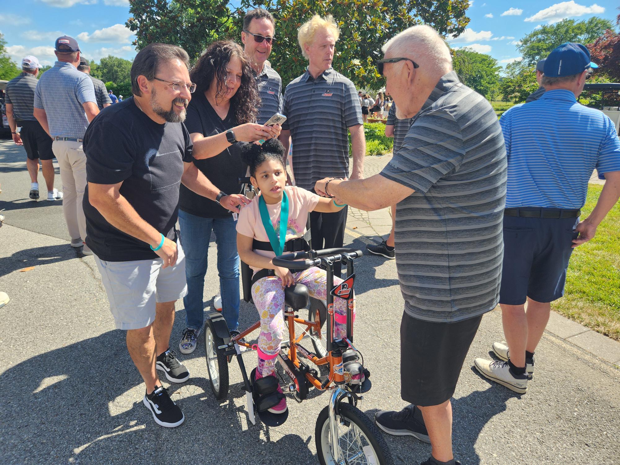 Flyers golf outing participants are outside with 8-year-old Josie Johnson on her new Flyers themed orange and black adaptive bike from Freedom Concepts. Josie has light brown skin and black hair in afro puffs.