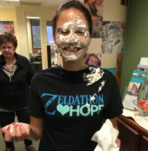 Director of Communications for Help Hope Live Shannon Shensky has a face covered in pie and a Zeldathon Hope t-shirt as she stands in the Help Hope Live headquarters. She has light skin and brown hair.