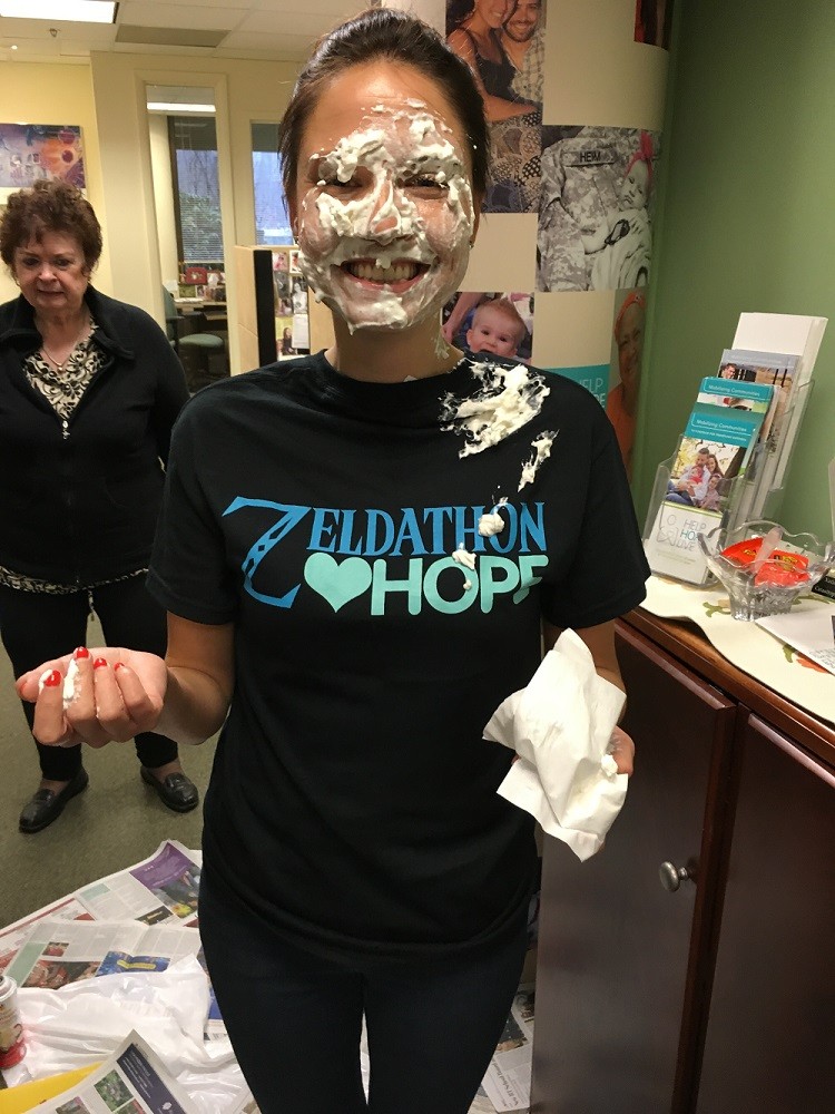 Director of Communications for Help Hope Live Shannon Shensky has a face covered in pie and a Zeldathon Hope t-shirt as she stands in the Help Hope Live headquarters. She has light skin and brown hair.