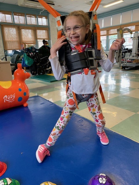 12-year-old Sabina Heim in a therapy harness. She has light skin, glasses, and blonde hair.