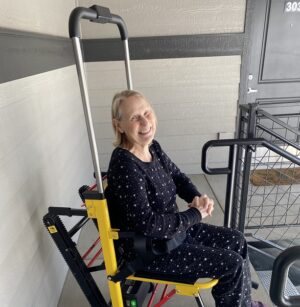 Kristina "Kristy" Cosden is pictured seated in a therapeutic wheelchair device that is yellow with a large silver grab bar above it. Kristina has light skin and shoulder-length gray hair and wears comfortable navy clothing with white dots.