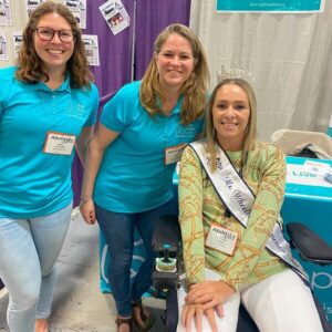 Kate and Sonny are in front of the Help Hope Live booth of Abilities Expo NY Metro with client and Ms Wheelchair America Ali Ingersoll. Kate has light skin, curly brown hair, and glasses and wears a Help Hope Live teal polo. Sonny has sandy hair just past her shoulders, light skin, and a matching polo. Ali Ingersoll has light skin, blonde hair just past her shoulders, and a sparkling tiara with a Ms Wheelchair America sash across her chest. She is seated in a black power chair.