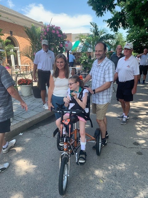 Sabina and her family members outside at the Flyers Alumni golf outing as Sabina sits on her new Flyers themed adaptive bike.