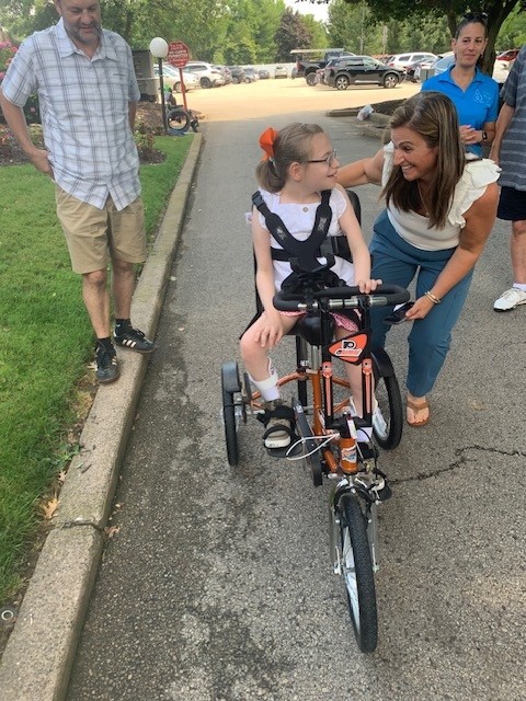 12-year-old Sabina smiles at her mother on her new Flyers themed adaptive bike. She has light skin, sandy hair, and glasses.