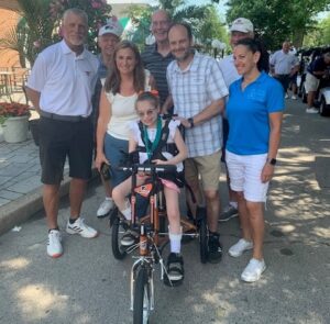 12-year-old Sabina surrounded by family, Flyers Alumni, and Help Hope Live's Kelly L Green outdoors as she sits on her new adaptive bike.