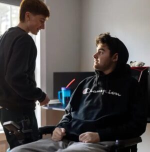 20-year-old Jack Weeks is seated in his power chair speaking with a standing friend at home.