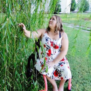 Help Hope Live client Jamie Carlson aka the "Cerebral Palsy Quilter" is outside in her manual wheelchair reaching for the branches of a weeping willow.