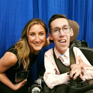 Sarah Burcaw with cousin Shane Burcaw, co-founders of Laughing At My Nightmare. Sarah holds an award and Shane is seated in his power chair.