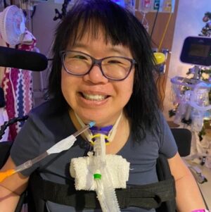 Kate Kosaka after her spinal cord injury in a hospital smiling with a ventilator tube seated in a wheelchair.
