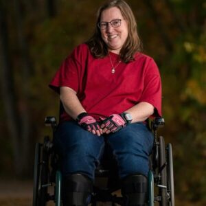 A professional portrait photo of Nicole Sanders outdoors seated in her manual wheelchair with leg braces and wheelchair gloves.