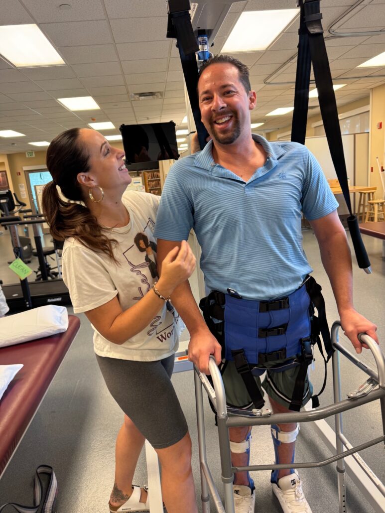 Shawna stands beside Jason Ellefson as he grins and stands with assistance in rehabilitation.