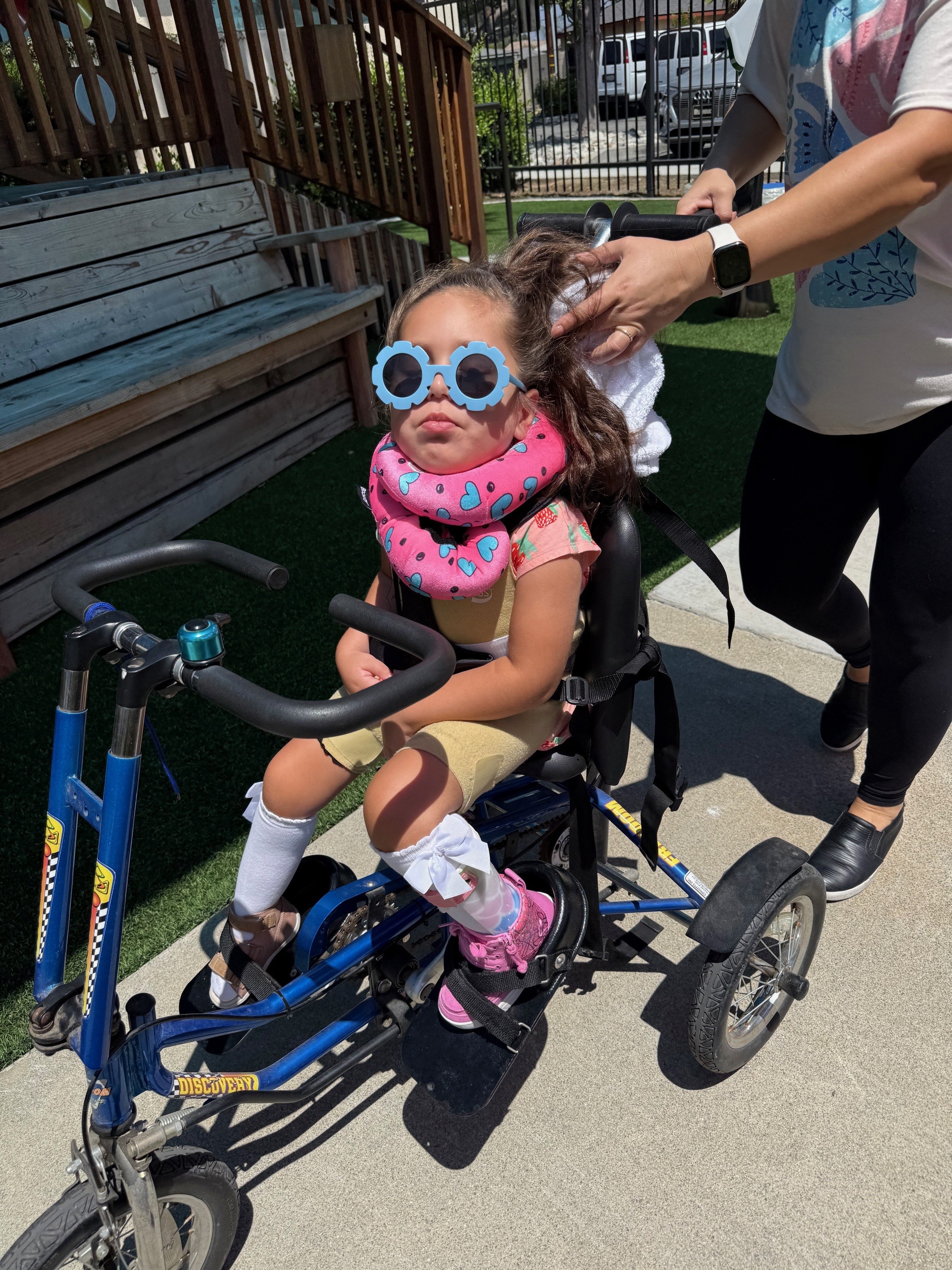 Brynlee Bonilla rides her adaptive bike outdoors with assistance.