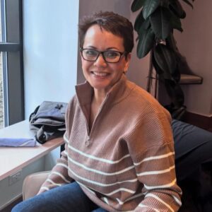 Pictured prior to a 2025 aneurysm, Jovita McConnell is seated in a chair wearing glasses with short red-brown hair.