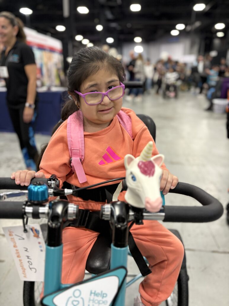 Jeanney sits on her new adaptive bike, which is teal with a unicorn at the front, at Abilities Expo NY 2026.