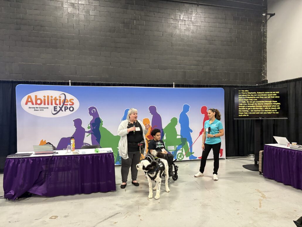 Stephanie Williams speaks about Help Hope Live with Kelson Williams, seated in his manual wheelchair, and service dog Gus alongside Kelly L Green at Abilities Expo NY 2026.