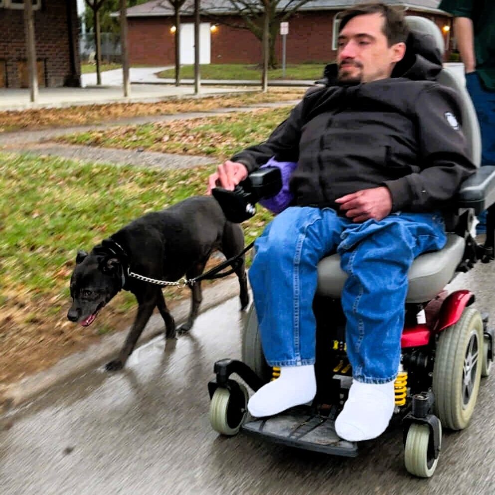 Scott Hughes seated in a power chair rolling down the street walking dog Karma.