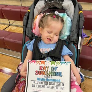 Kaylie is seated in her stroller with her Ray of Sunshine award from her school.