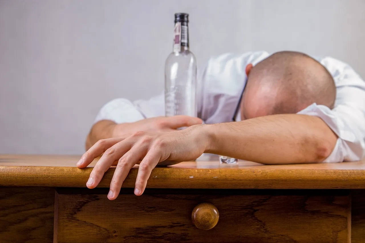 A person resting their head on a desk with an empty bottle of alcohol next to them