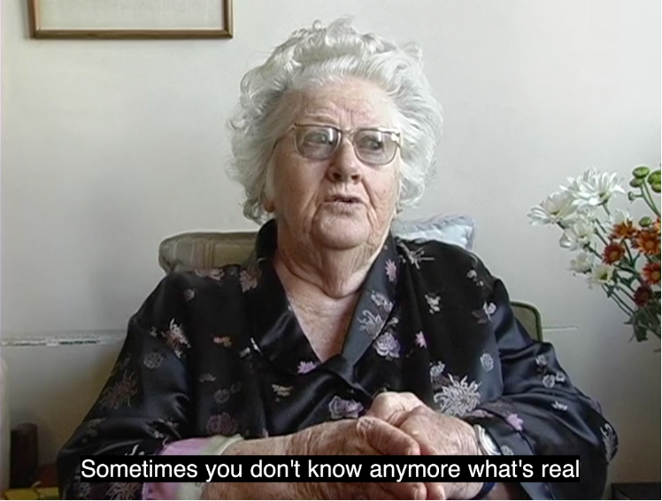 A movie still showing an older woman with white hair and glasses, wearing a black flowery blouse. Off-screen there are flowers and a picture frame.
The woman appears to be talking, she is gesticulating. The closed caption reads: 'Sometimes you don't know anymore what's real' 