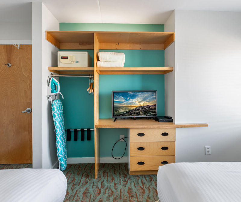 A hotel room with a desk, shelves, and a TV between two beds.