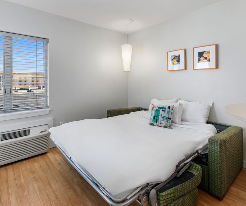 A small guest room with a sofa bed, window, air conditioner, and artwork
