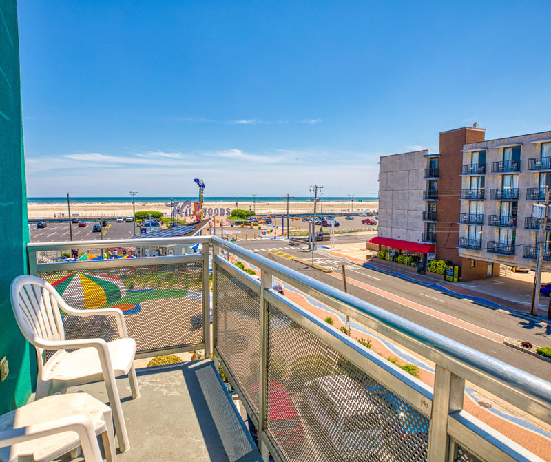 A balcony with a metal railing and white chairs overlooking a beach and street.