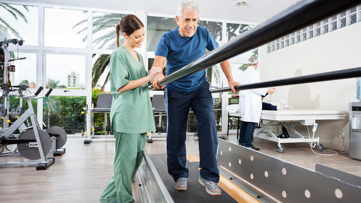 Female physiotherapist standing by smiling senior patient walking between parallel bars in rehab center