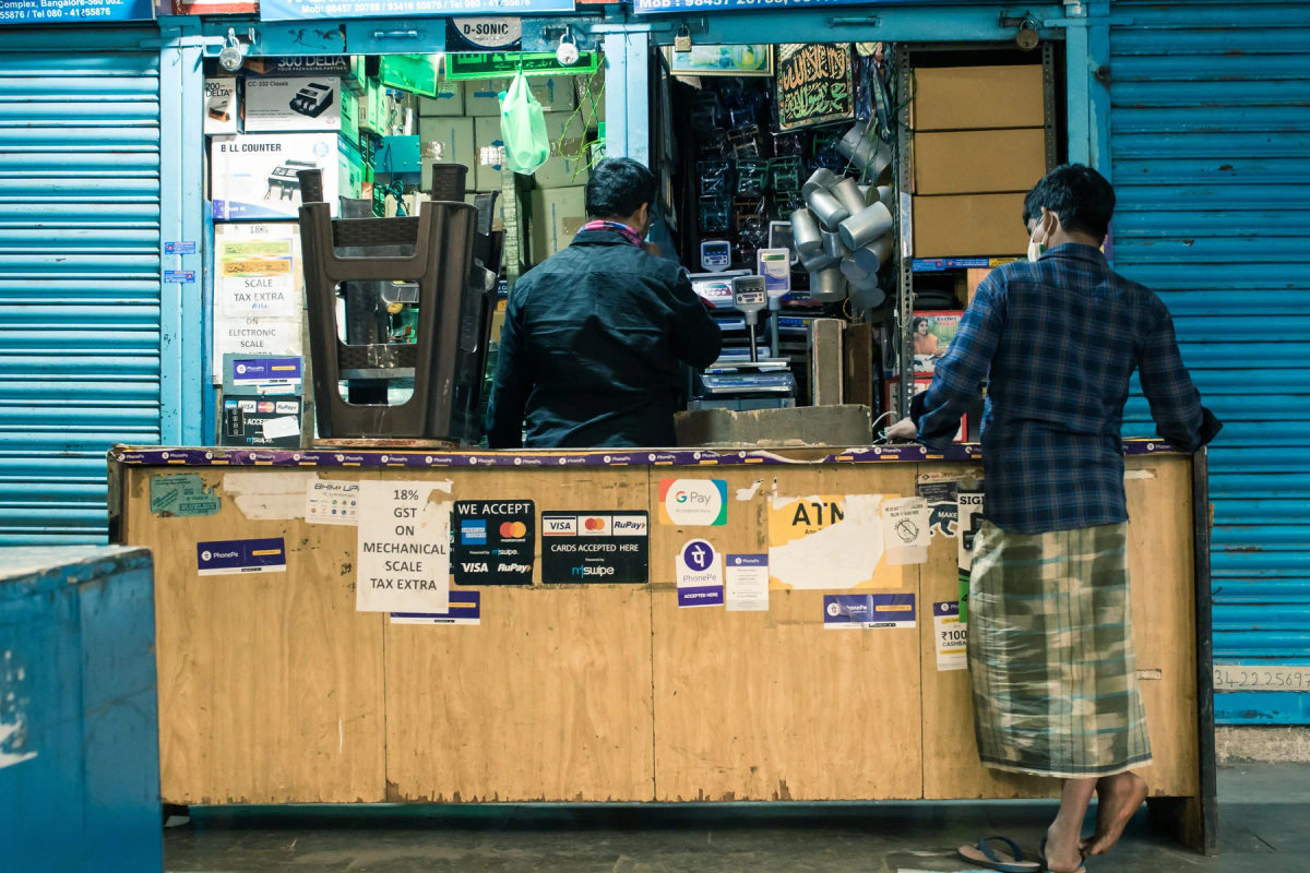 Two men stand at a shop counter in a Bengaluru marketplace with payment signs and products displayed.