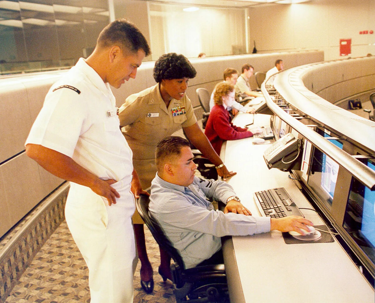 A technician works at a desktop computer in a shipboard setting.