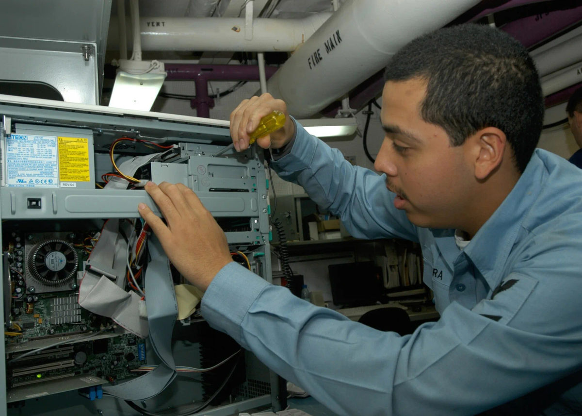 A technician works at a desktop computer in a shipboard setting.